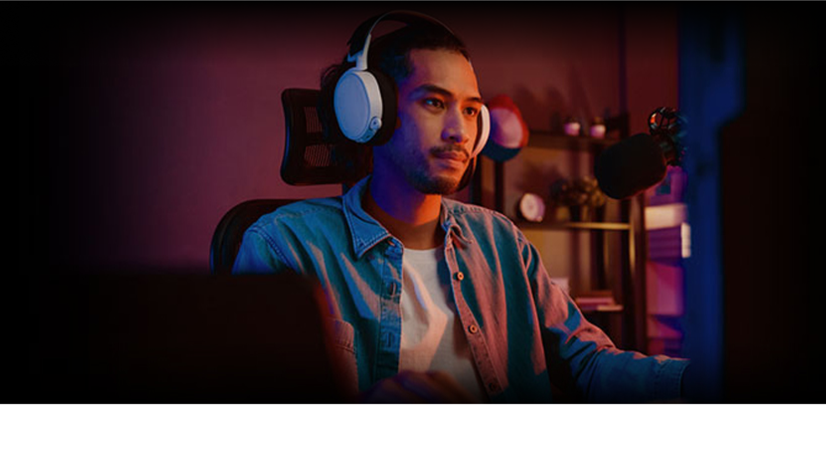 A man wearing headphones sits at a desk with a computer and microphone, focused on the screen. The room is dimly lit with purple and orange lighting, and shelves with various items are visible in the background.