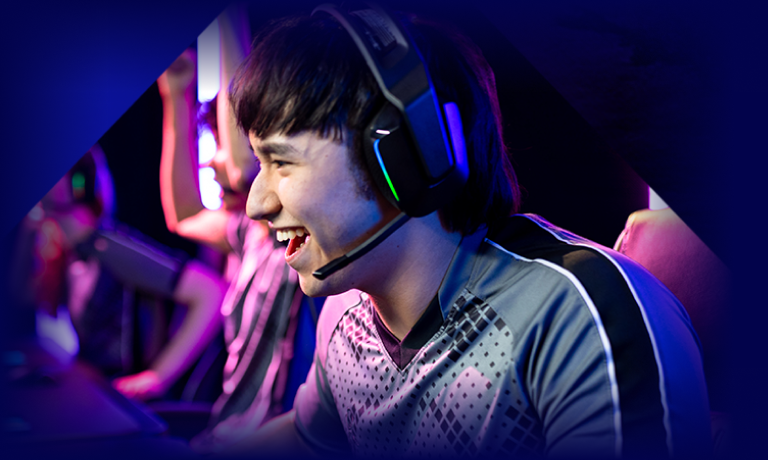 A young person wearing a headset and a gaming jersey smiles excitedly while playing a video game in a brightly lit gaming environment. Other teammates are visible in the background.