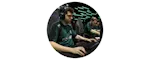 Two people wearing dark green jerseys and headsets are seated at computers, focused on gaming in a competitive or team environment.