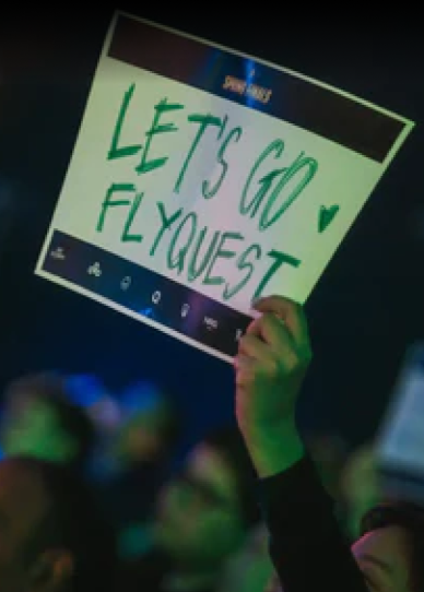 A person holds up a sign that reads LETS GO FLYQUEST with a small heart, in a crowd at an event.